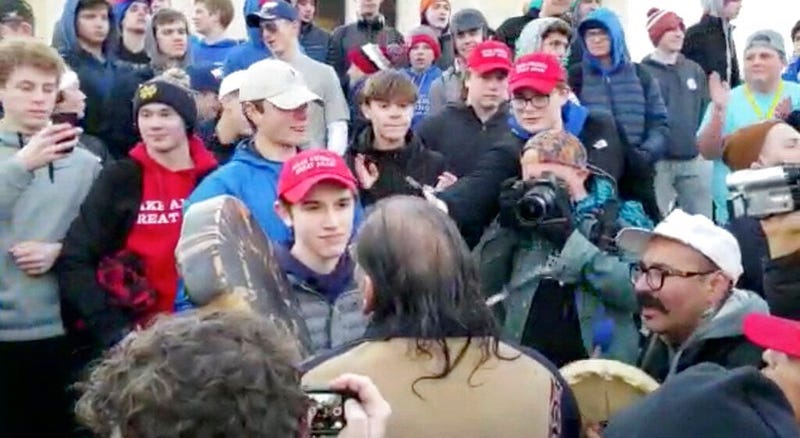 In this Friday, Jan. 18, 2019 image made from video provided by the Survival Media Agency, a teenager wearing a "Make America Great Again" hat, center left, stands in front of an elderly Native American singing and playing a drum in Washington. 