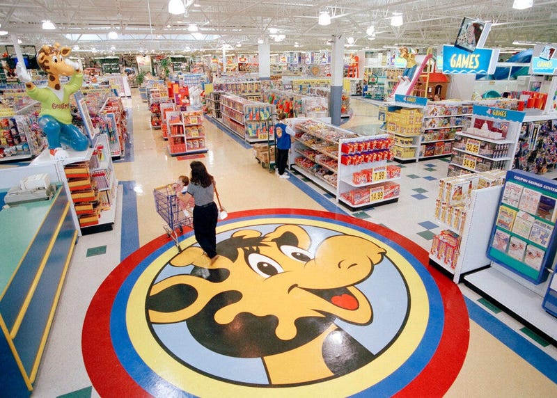 a woman pushes a shopping cart over a graphic of Toys R Us mascot Geoffrey the giraffe at the Toys R Us store