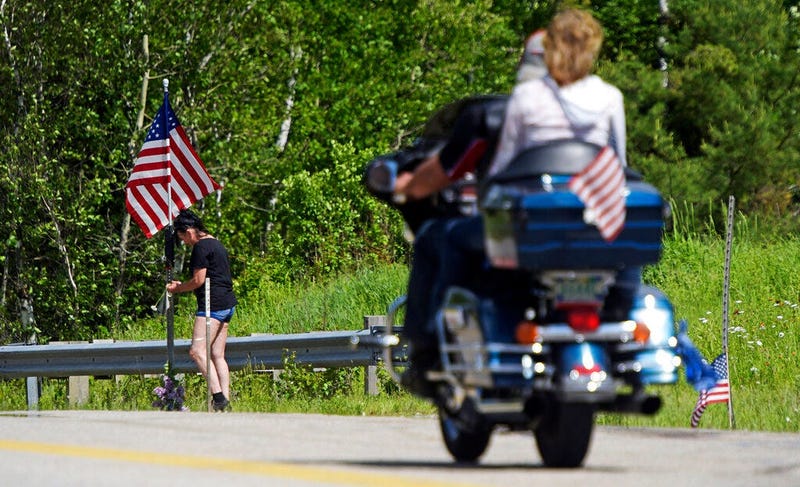 A motorcycle passes as a woman leaves flowers at the scene of a fatal accident on Route 2 in Randolph, N.H., Saturday, June 22, 2019. Investigators pleaded Saturday for members of the public to come forward with information that could help them determine