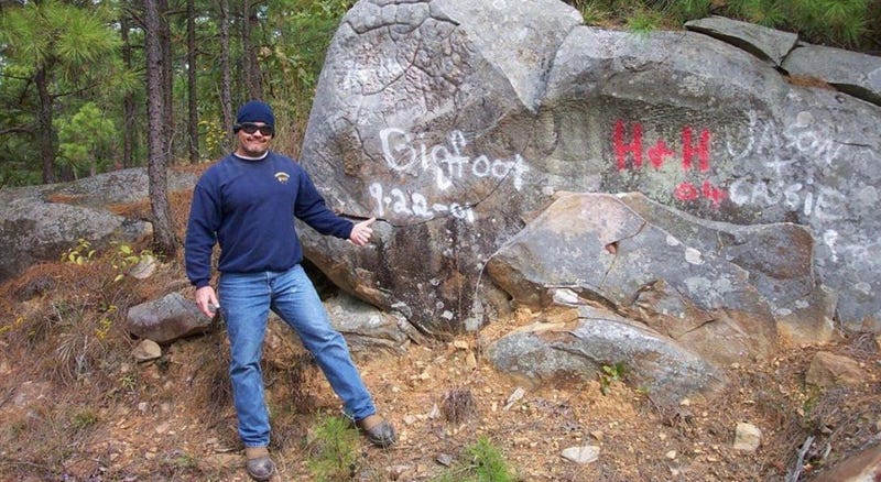 Randy Harrington, Bigfoot enthusiast, poses next to a rock w the word 'Bigfoot" spray painted on it