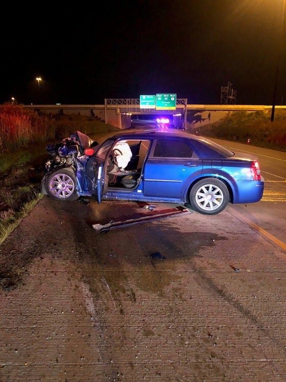 A blue 2009 Chrysler 300 traveling westbound drove onto the inside shoulder and struck the rear of the parked police car at a high rate of speed.