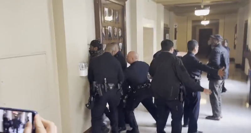 several police officers handcuff a man against the wall outside the council chamber