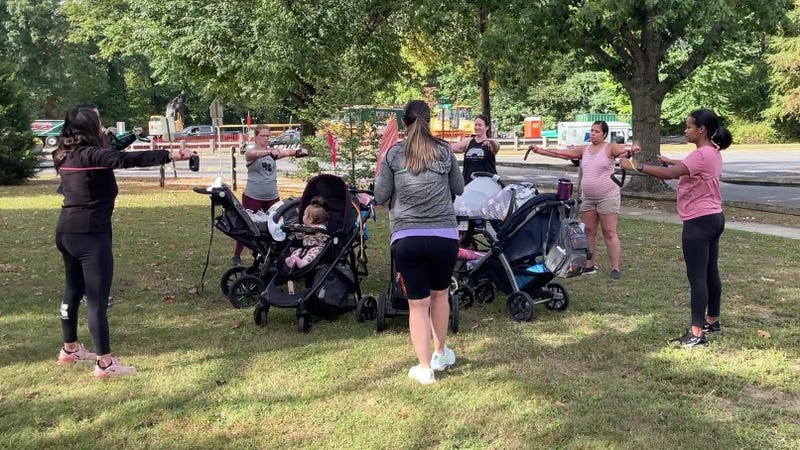 A group of mothers exercise in a park while their children in strollers look on.