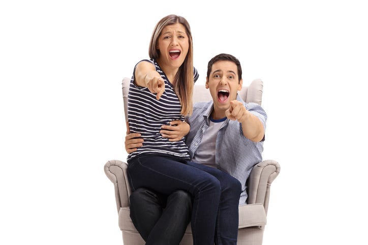 Happy young couple in an armchair pointing at the camera and laughing isolated on white background