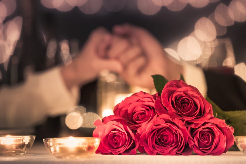 Couple at a candle light dinner date holding hands next to bouquet of red roses.