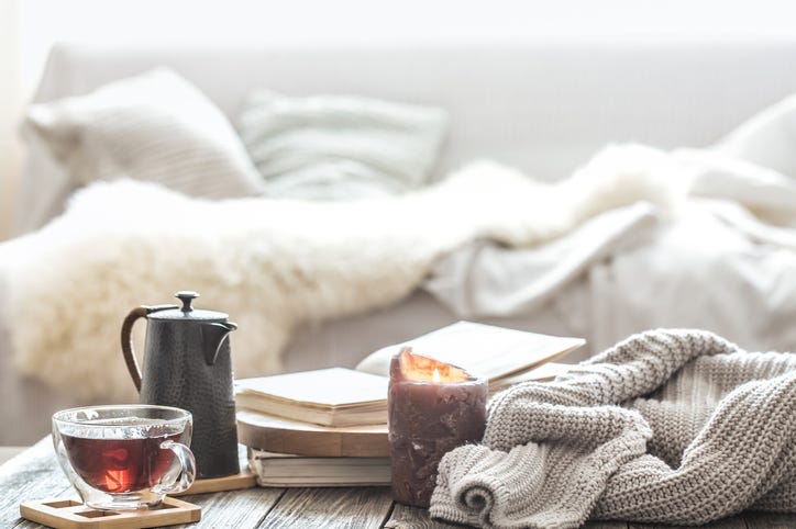A cup of tea and a kettle, near a burning candle and a knitted sweater.