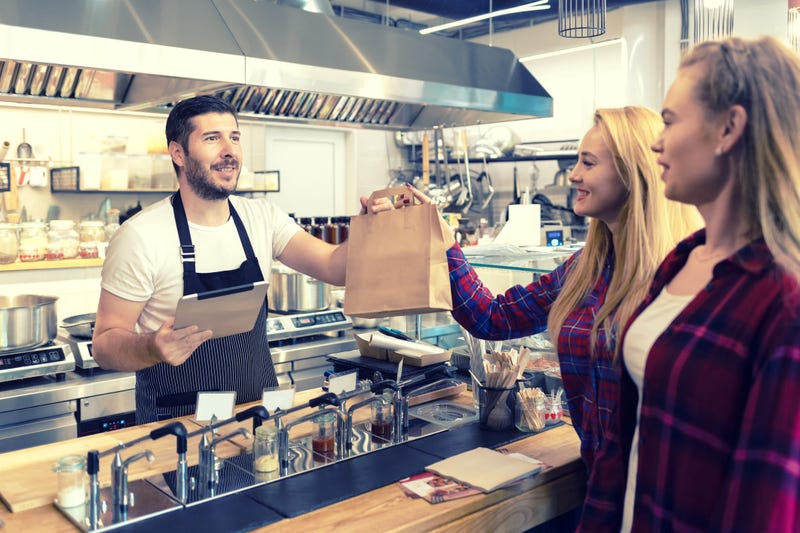 Waiter serving takeaway food to customers at counter in small family eatery restaurant - trendy fast food smiling owner delivering an online to go order in recycled paper bag to clients - stock photo