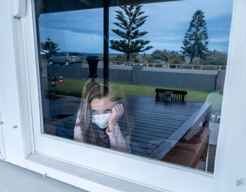 Depressed lonely little girl with face mask looking through the window during quarantine.
