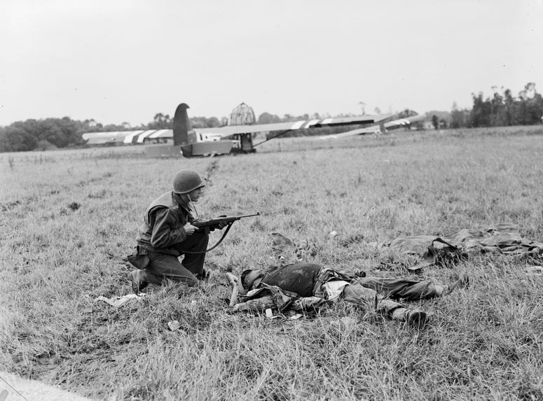 An American soldier crouching by the body of a dead German soldier with US Waco troop carrying gliders on the field behind him in Normandy.