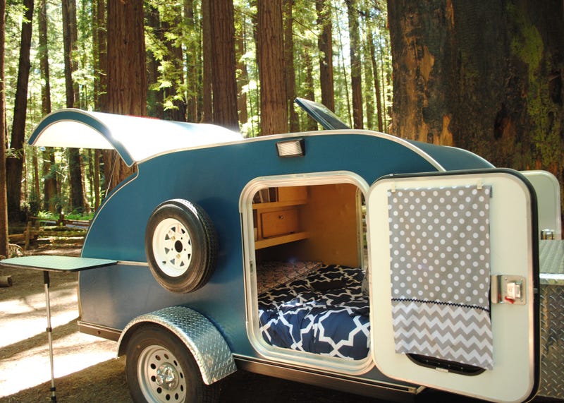 Blue teardrop camping trailer with doors open and view to interior, setup at a campsite surrounded by redwood trees in Humboldt Redwoods State Park in California.