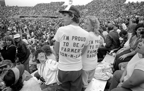 View of attendees at the debut Farm Aid benefit festival, Champaign, Illinois, September 22, 1985. Two stand wearing t-shirts with matching slogans, 'I'm Proud to be a Farmer's Son-in-Law' and 'I'm Proud to be a Farmer's Daughter.' 
