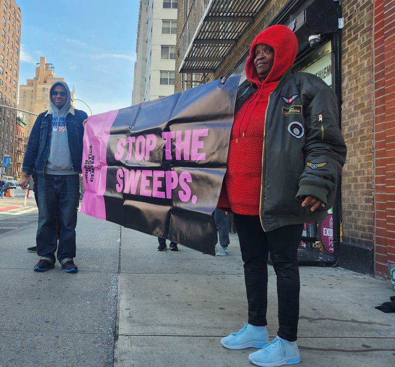 Crystal Vails (right) and a worker for the Urban Justice Center's Safety Net Project (left) hold a sign reading "stop the sweeps" as NYPD officers, sanitation workers and Department of Homeless Services workers approach to force Vails to move her belongings.