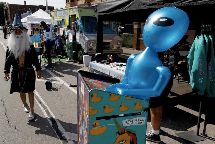 Photo of a booth at Dally in the Alley with a giant inflatable alien set in a box with rubber duckies and a unicorn printed on the side and "...transcend the law"; a person dressed like a wizard walks in the direction towards the camera; a truck/van is also scene in the background