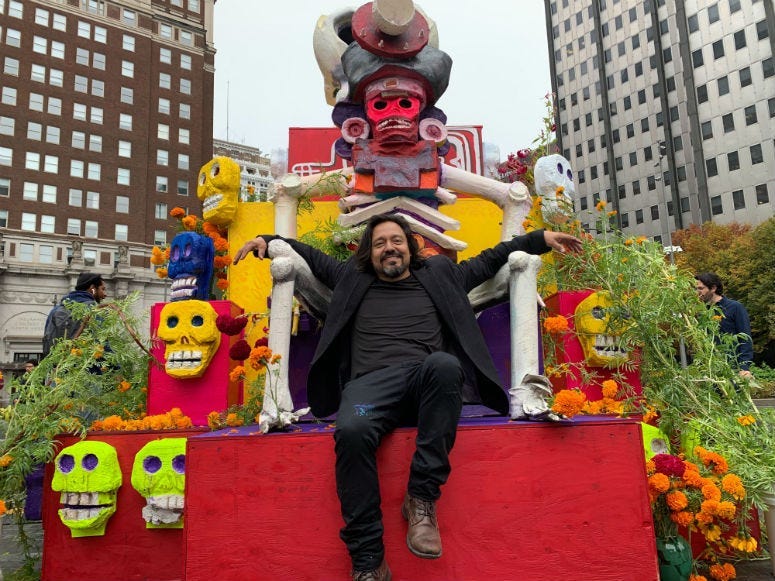 Artist Cesar Vivaros sitting on top of the Dia De Muertos altar he created that in Love Park.