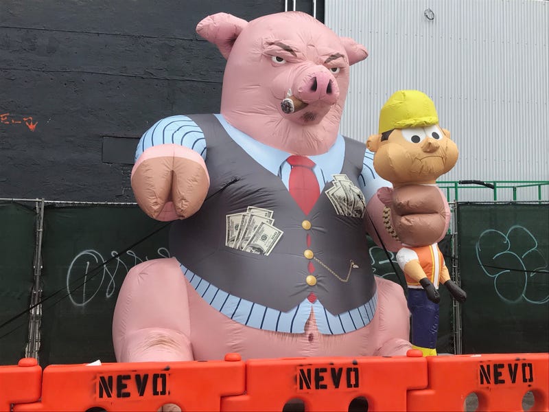 An inflatable pig has a Rolex on his wrist, pockets full of cash and a cigar in his mouth, and one of his hooves is wrapped around the neck of a construction worker.