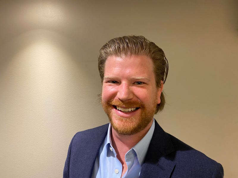 Jack Coatar stands smiling. he has dirty blond hair and beard and blue suit on
