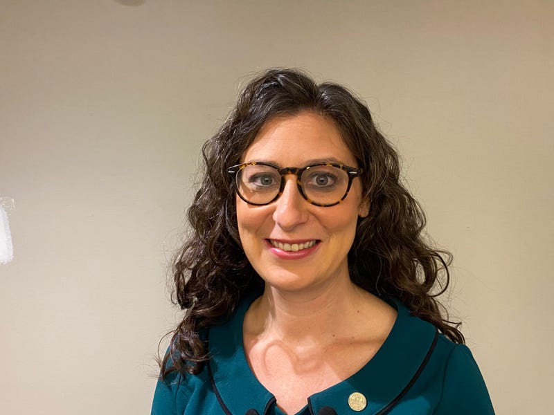 megan green stands smiling. she has long curly brown hair and glasses and a blue shirt.