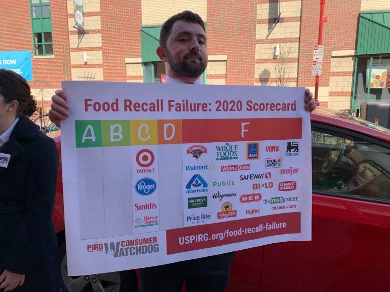 A man holds up a sign that has information on food recall failures by grocery stores. 
