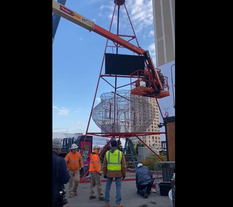 Orange crane in front of the world's largest Salvation Army Red Kettle installed in Cadillac Square, across from Campus Martius Park, in downtown Detroit; Detroit skyline and clouds are visible in background