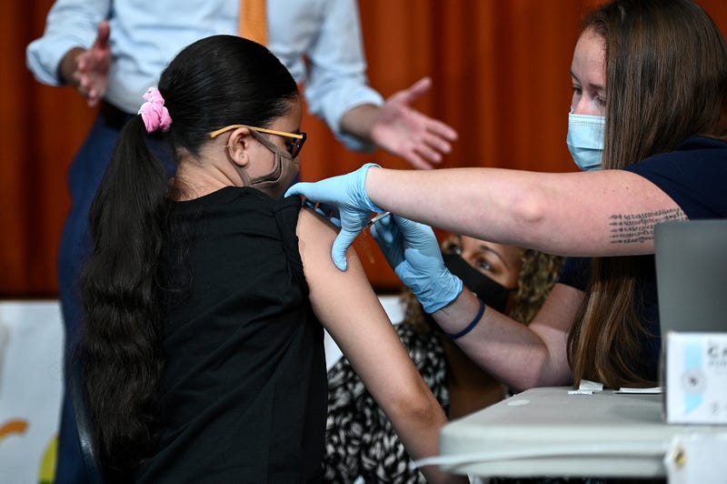 File photo: With New York City Mayor Bill de Blasio (orange tie) and New York City Schools Chancellor Meisha Porter (black mask) looking on, Giret Madina, 14, receives her first dose of the Pfizer BioNTech COVID-19 vaccine at a pop-up vaccine site located at the Lehman High School Campus in the Bronx on July 27, 2021