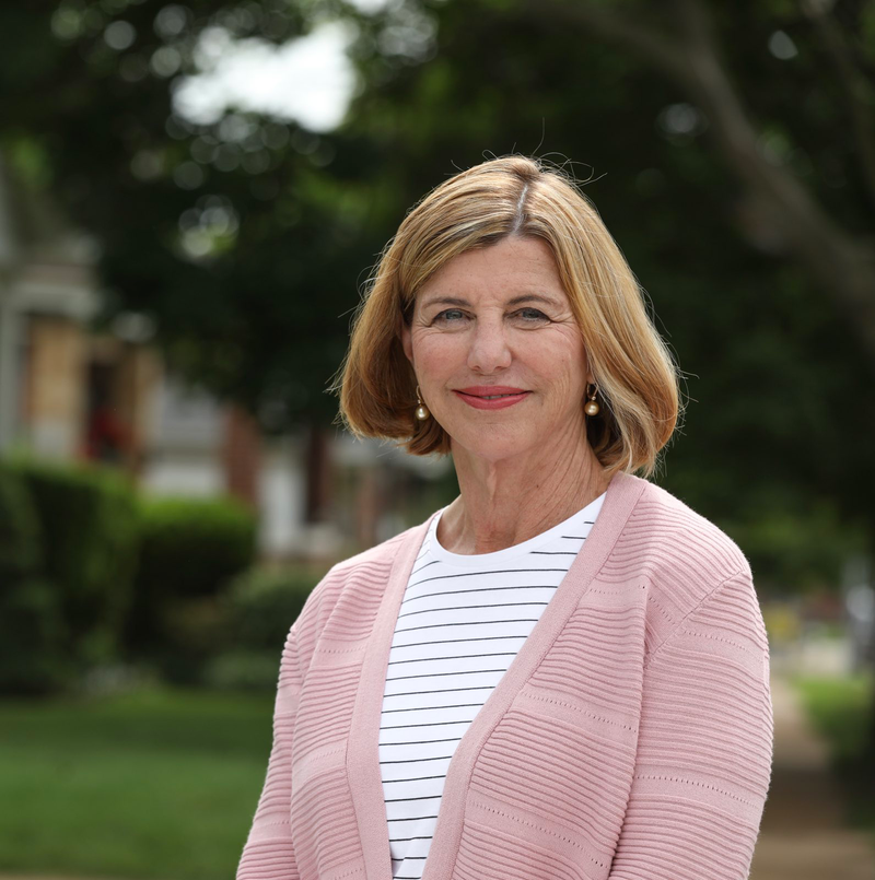 Trudy Busch Valentine stands outside smiling. she is a white woman with short strawberry blond hair, a black and white striped t shirt and a light pink cardigan