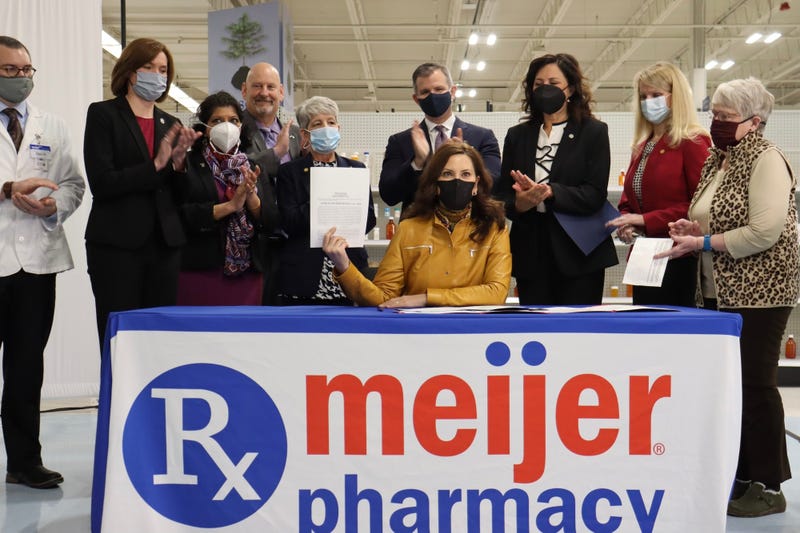 Gretchen Whitmer holding up signed bill sitting at table that says Meijer pharmacy and surrounded by people standing and clapping